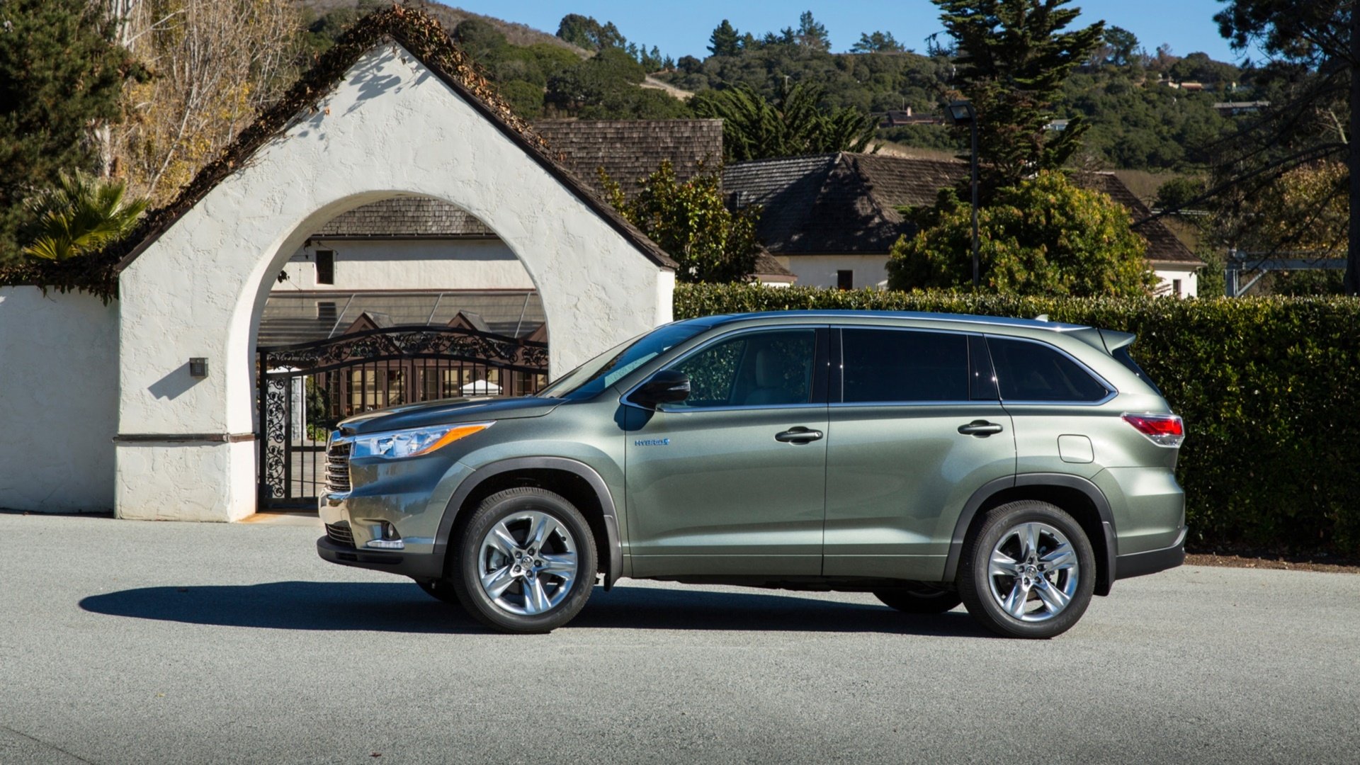HD desktop wallpaper featuring a Toyota Highlander Hybrid parked in front of a white arched gate with greenery and cottages in the background.