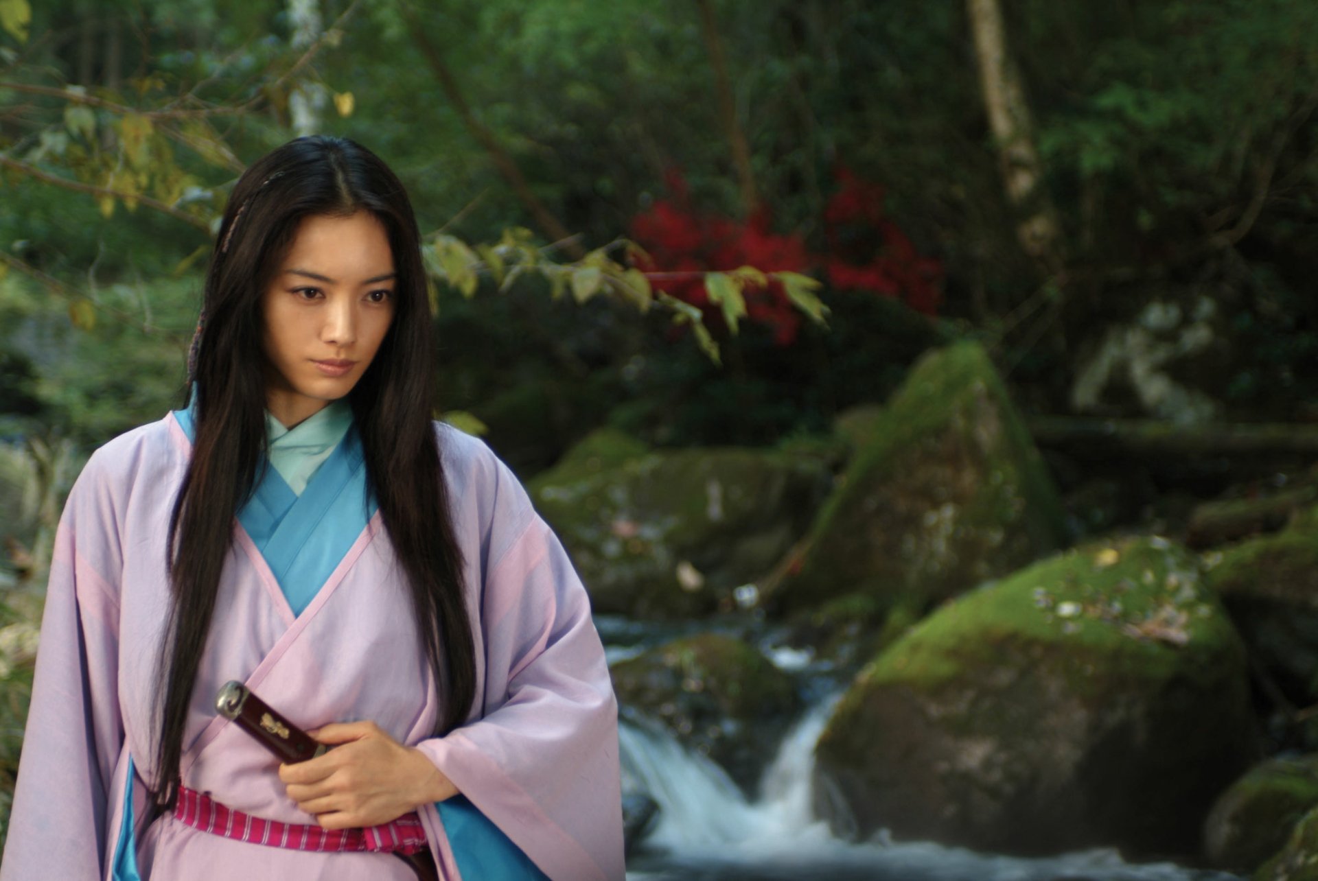 HD PC desktop wallpaper from the movie Shinobi: Heart Under Blade — a female shinobi in a pink kimono clutching a sheathed blade beside a mossy forest stream.