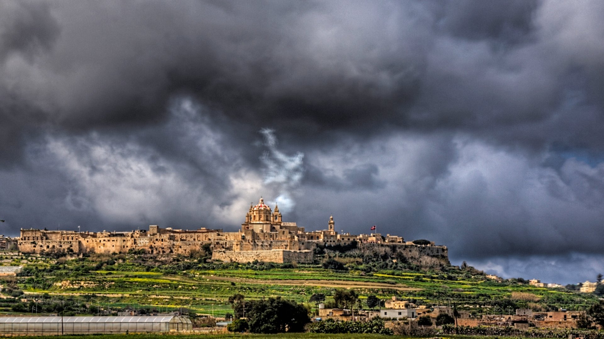 HD desktop wallpaper featuring the man-made historic cityscape of Mdina under a dramatic, stormy sky.