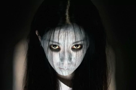 HD desktop wallpaper of the pale-faced, long-haired ghost from Ju-on: The Grudge, hollow dark eyes and stringy black hair framed by shadowed, eerie background.