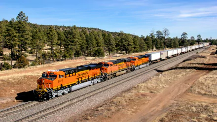HD PC desktop wallpaper featuring a vibrant orange BNSF freight train traveling through a forested landscape under a clear blue sky.