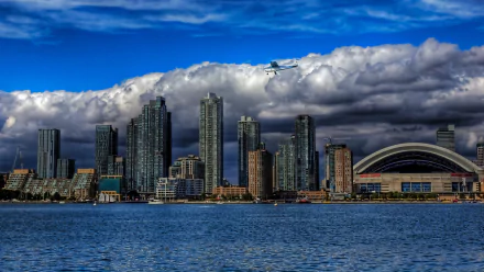 A stunning HD desktop wallpaper showcasing Toronto's skyline, featuring towering buildings against a dramatic sky with clouds and an airplane flying overhead.