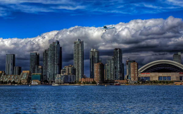 A stunning HD desktop wallpaper showcasing Toronto's skyline, featuring towering buildings against a dramatic sky with clouds and an airplane flying overhead.