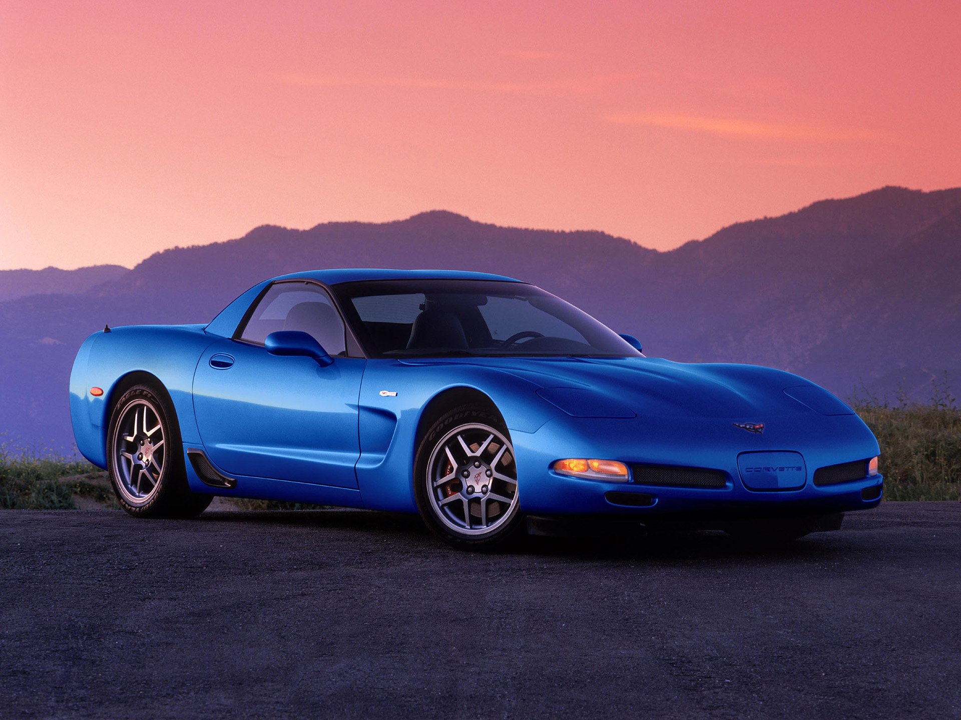 A blue Chevrolet Corvette Z06 parked on a road with mountains and a pink-orange sunset sky in the background, shown as an HD PC desktop wallpaper.