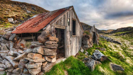 HD PC desktop wallpaper and background: man-made stone-and-wood cabin with a rusted corrugated roof nestled among rocky, grassy hills under a dramatic cloudy sky.