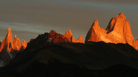 HD PC desktop wallpaper: Mount Fitz Roy and serrated peaks bathed in warm alpenglow above shadowed ridgelines under a dusky sky — nature landscape background.