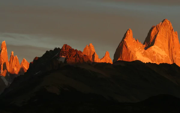 HD PC desktop wallpaper: Mount Fitz Roy and serrated peaks bathed in warm alpenglow above shadowed ridgelines under a dusky sky — nature landscape background.