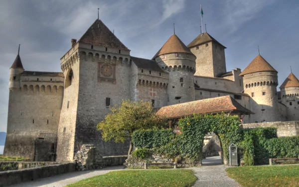 HD wallpaper of Château de Chillon, a historic man-made castle in Veytaux, Switzerland, featuring stone towers and battlements under a cloudy sky.
