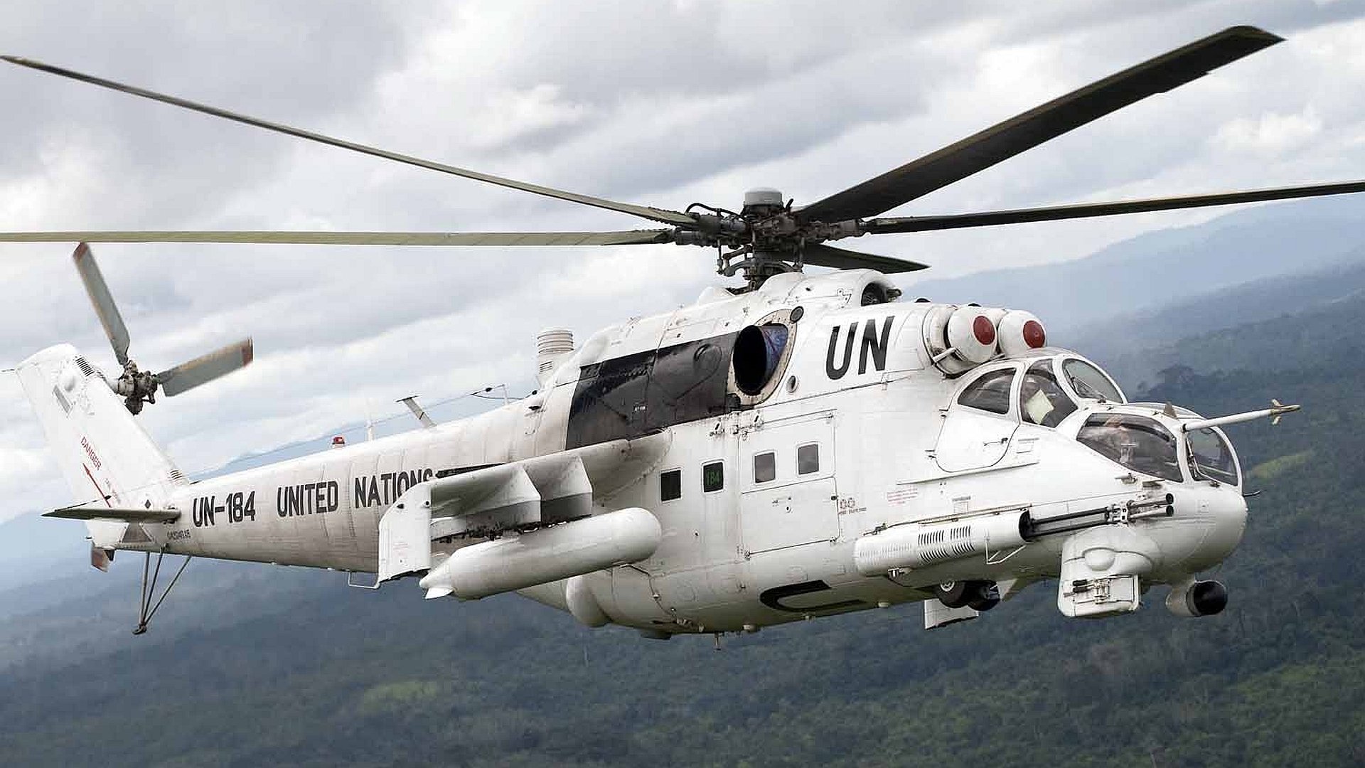 HD PC desktop wallpaper: white United Nations Mil Mi-24 attack helicopter in flight over mountains, military rotorcraft against a cloudy sky