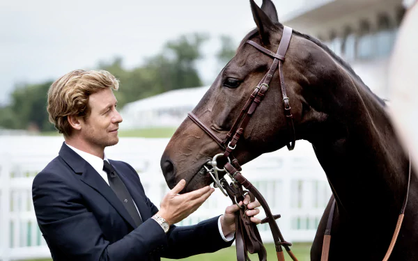 Australian actor and celebrity Simon Baker, dressed in a suit, gently interacts with a dark brown horse in an outdoor setting, captured in a high-definition desktop wallpaper.