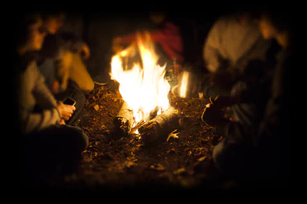 HD desktop wallpaper showing a group of people gathered around a bright campfire at night, capturing the warm glow and cozy atmosphere of camping.