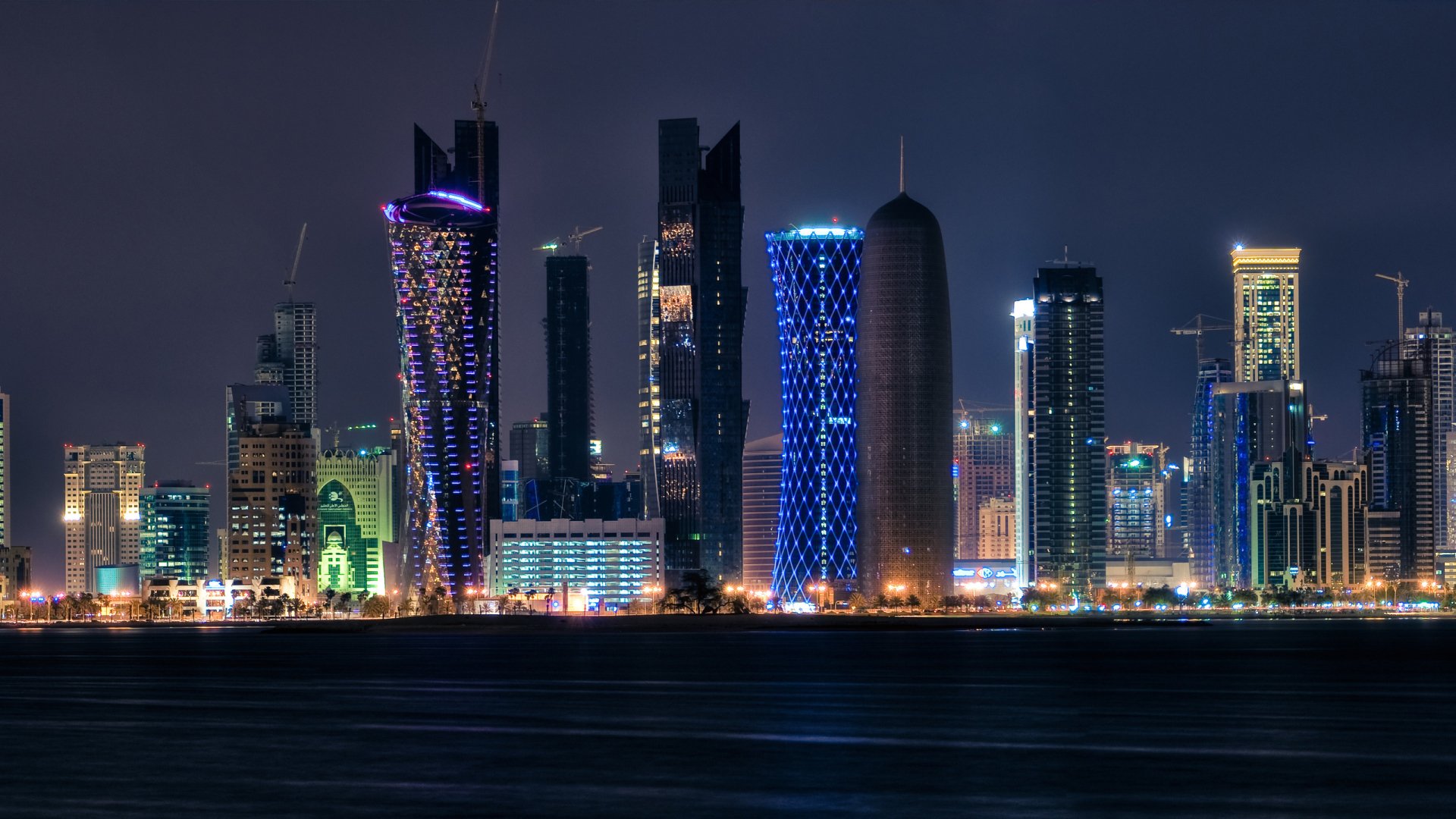HD desktop wallpaper of Doha's skyline at night, featuring illuminated, man-made skyscrapers against a dark sky, reflecting vibrant colors.