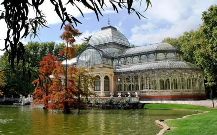 HD PC desktop wallpaper of the man-made Palacio de Cristal: a glass conservatory by a tranquil pond, framed by green and autumnal trees under a blue sky.
