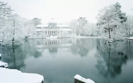HD desktop wallpaper of the Palacio de Cristal surrounded by snow-covered trees, reflecting on the calm water in a serene winter setting.