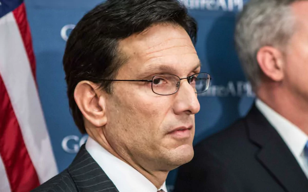 HD desktop wallpaper of a man in a suit with glasses looking to the side with an American flag in the background.