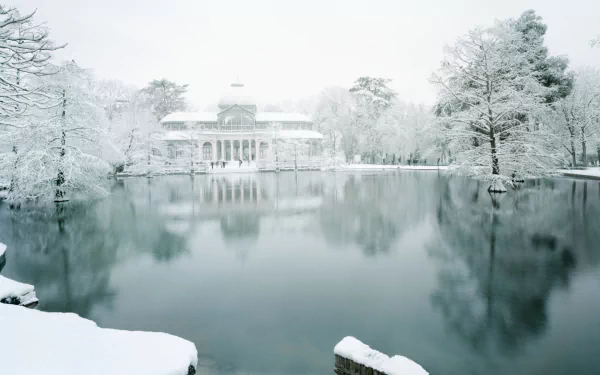 HD desktop wallpaper of the Palacio de Cristal surrounded by snow-covered trees, reflecting on the calm water in a serene winter setting.