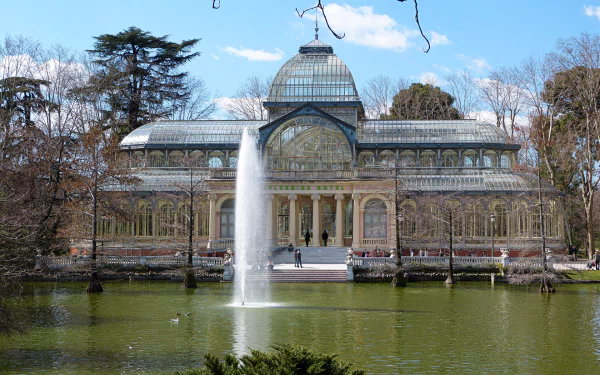 HD desktop wallpaper featuring the man-made Palacio de Cristal with a central fountain, surrounded by trees and a reflective pond under a clear blue sky.