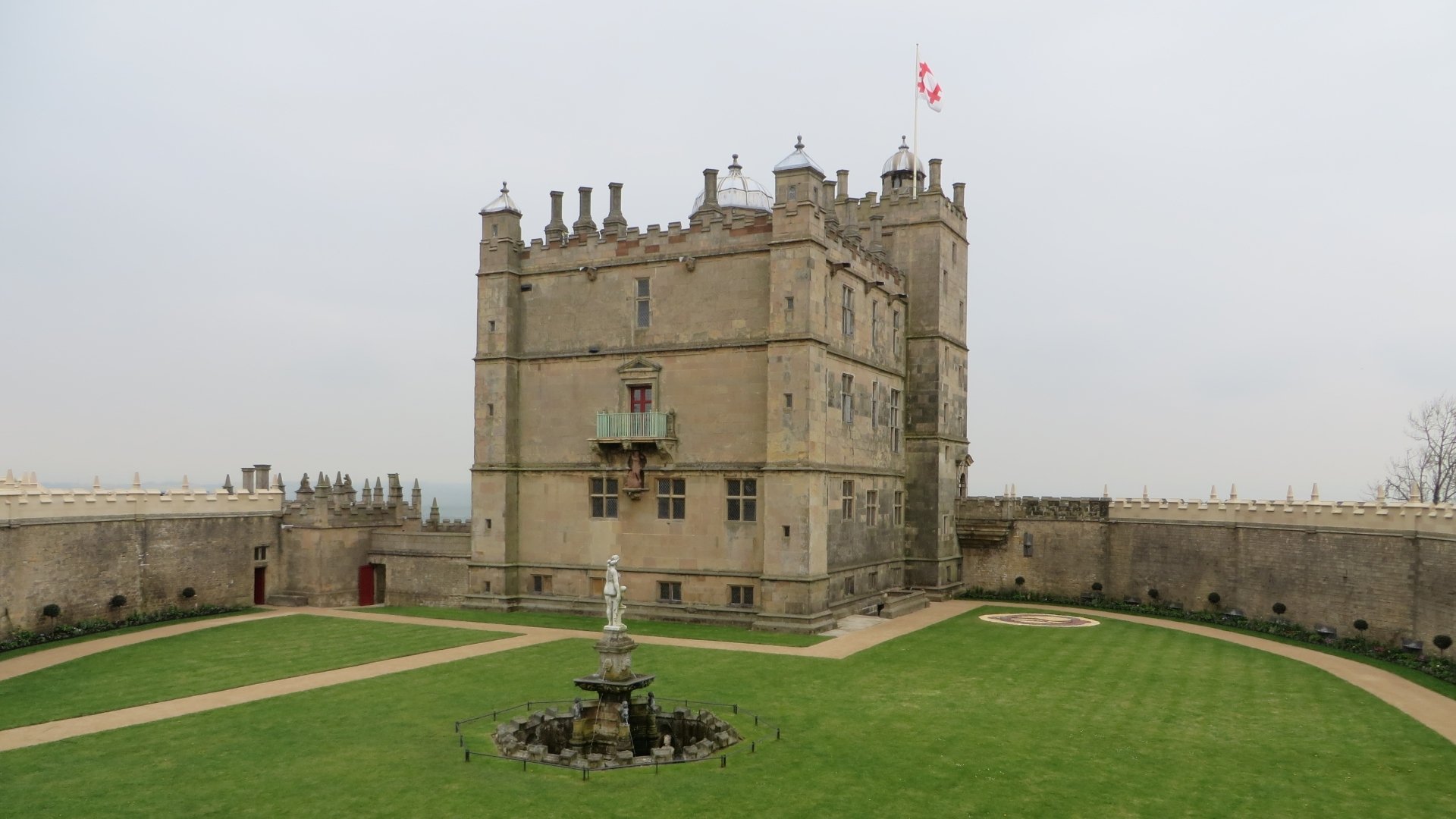 HD PC desktop wallpaper background of man-made Bolsover Castle: a stone central keep with flag, surrounding curtain wall and grassy courtyard with fountain under an overcast sky.
