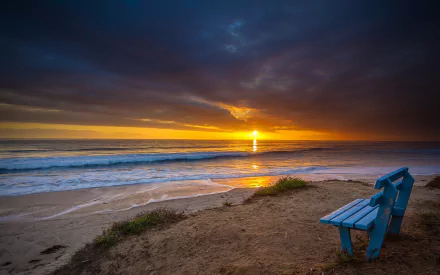 A solitary blue bench sits on a sandy beach, gazing out at a tranquil ocean under a dramatic sky, as the sun sets, creating a serene HD wallpaper.
