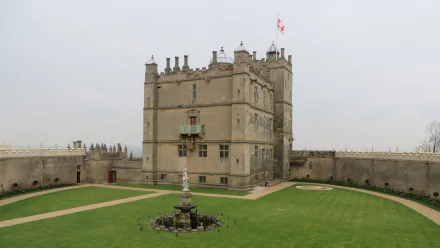 HD PC desktop wallpaper background of man-made Bolsover Castle: a stone central keep with flag, surrounding curtain wall and grassy courtyard with fountain under an overcast sky.