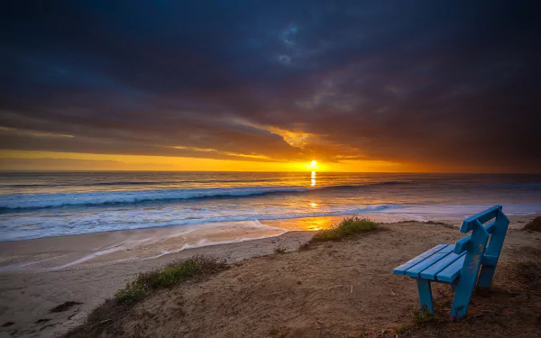 A solitary blue bench sits on a sandy beach, gazing out at a tranquil ocean under a dramatic sky, as the sun sets, creating a serene HD wallpaper.