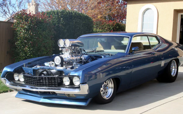 HD PC desktop wallpaper of a blue Ford Torino GT with a prominent supercharger, parked in a driveway against a backdrop of greenery and a house.