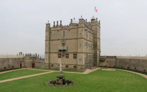 HD PC desktop wallpaper background of man-made Bolsover Castle: a stone central keep with flag, surrounding curtain wall and grassy courtyard with fountain under an overcast sky.