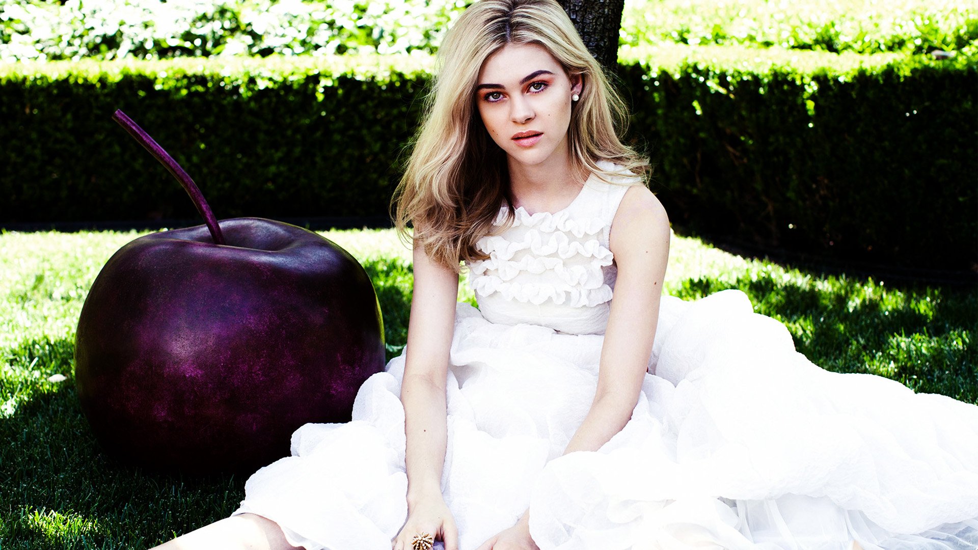 HD desktop wallpaper featuring celebrity Nicola Peltz seated outdoors in a white dress beside a large dark cherry, with lush greenery in the background.