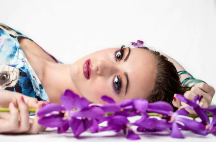 A woman with vibrant makeup lies against a white background, adorned with a cluster of purple flowers, creating a serene and striking HD desktop wallpaper.