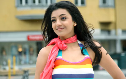A vibrant HD desktop wallpaper featuring Indian celebrity Kajal Aggarwal, smiling with confidence in a colorful outfit, set against a lively urban backdrop.