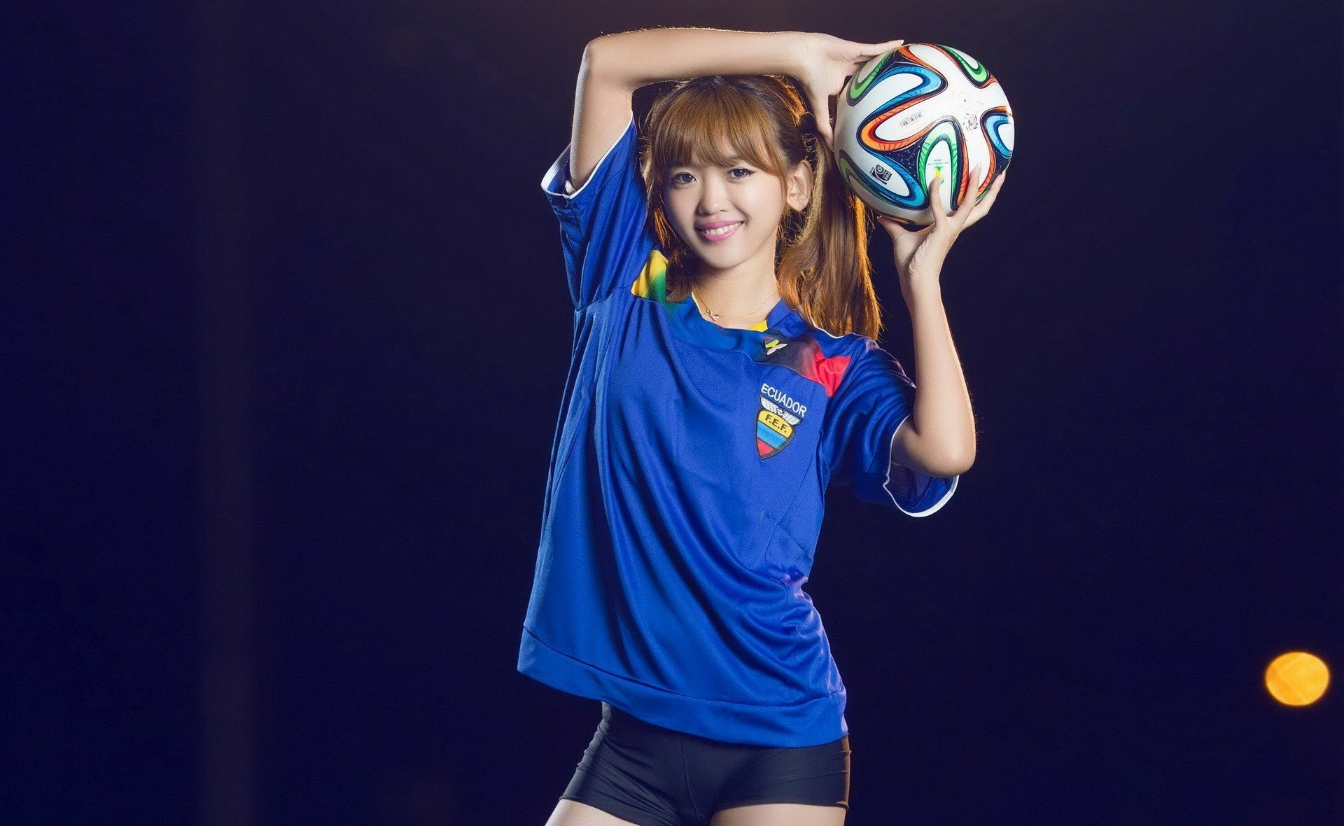 HD desktop wallpaper of an Asian woman in an Ecuador soccer jersey holding a soccer ball against a dark background.