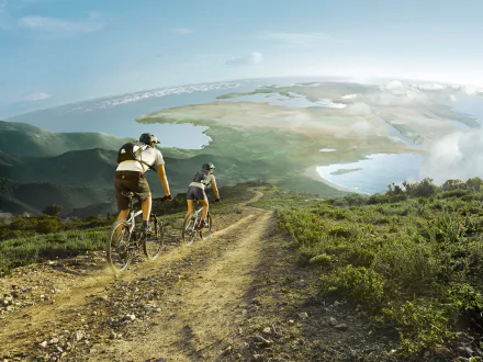 HD desktop wallpaper featuring two cyclists riding mountain bikes on a dirt trail overlooking a vast landscape with water and hills, capturing the spirit of outdoor sports.