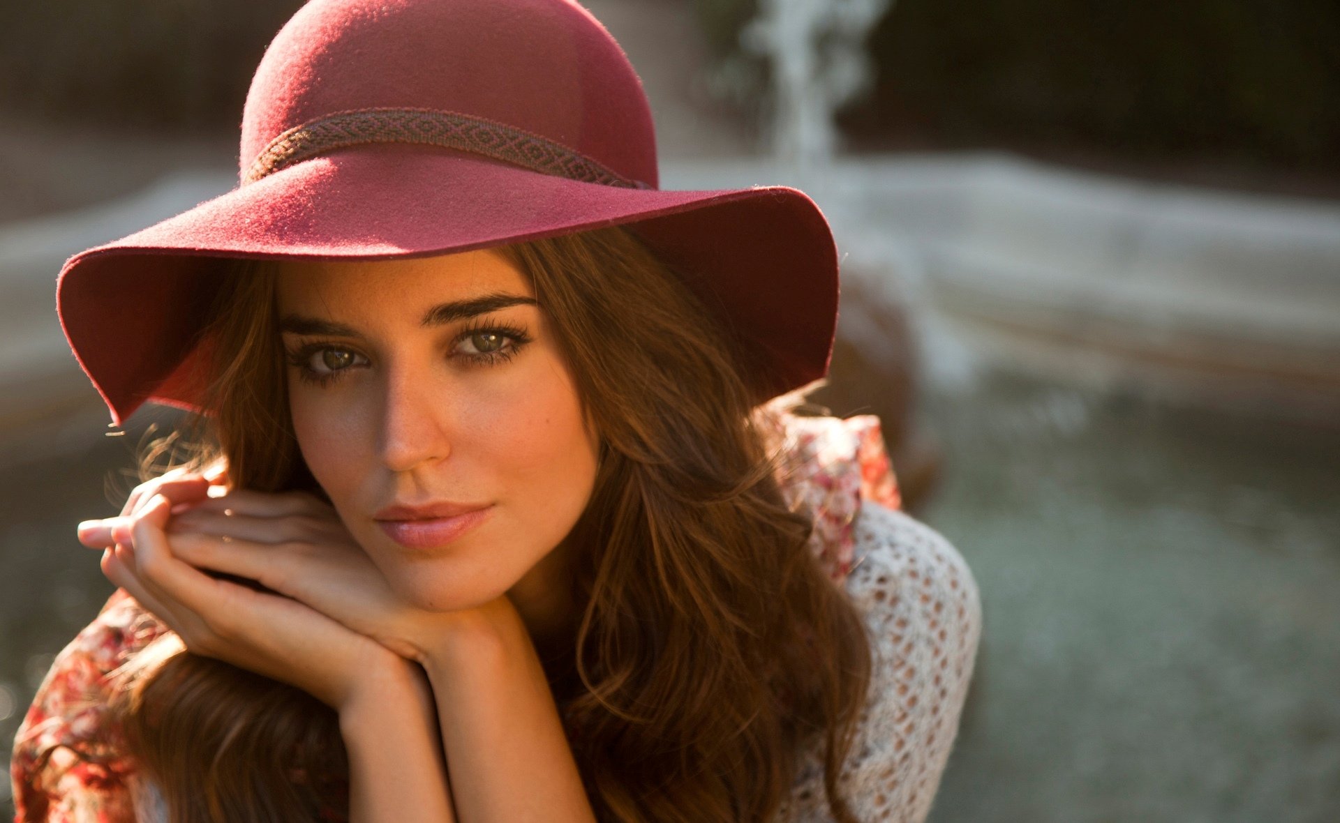 HD desktop wallpaper of Spanish model Clara Alonso wearing a wide-brimmed red hat, resting her chin on her hands with a blurred natural background.