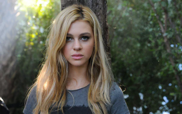 HD PC desktop wallpaper featuring American actress and celebrity Nicola Peltz with long blonde hair, standing outdoors against a blurred natural background.