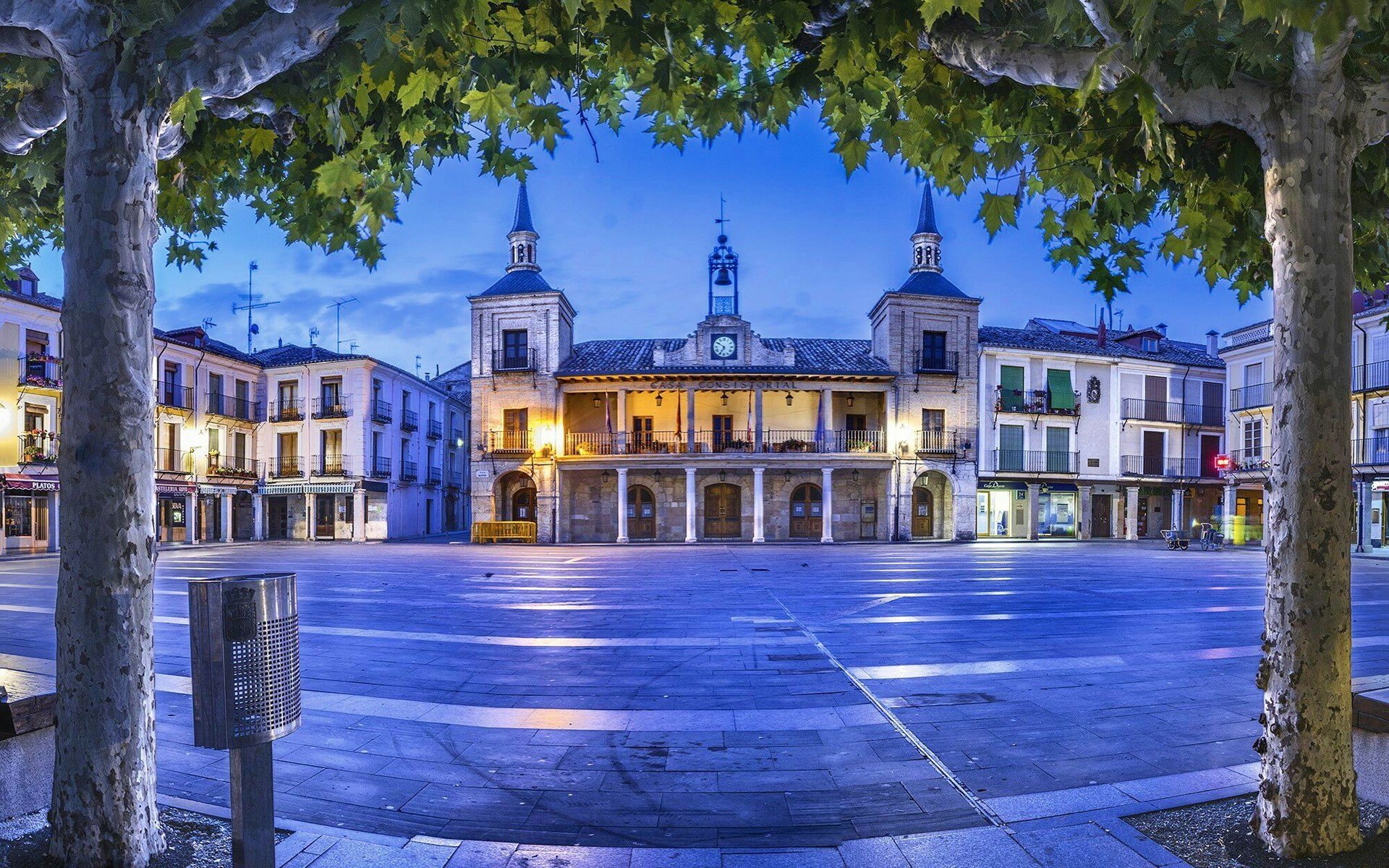 Religious church in a town square at dusk — HD PC desktop wallpaper and background showing illuminated bell towers, arched façade and a tree-framed plaza.