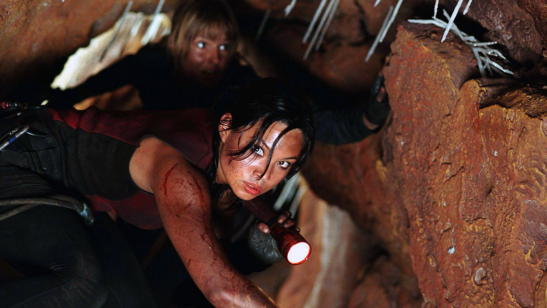 HD PC desktop wallpaper from the movie The Descent: blood-smeared woman crawls through a narrow cave with flashlight, stalactites overhead and a claustrophobic, tense horror atmosphere.