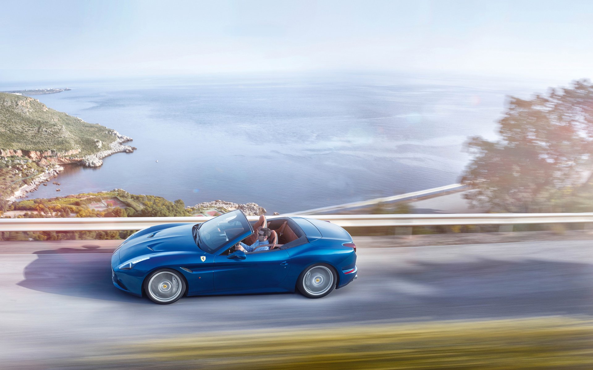 Blue Ferrari California T convertible cruising a coastal road with motion blur and sea view — 2K Quad HD PC desktop wallpaper/background.