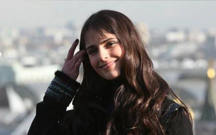 Smiling brunette Brazilian actress and celebrity in a sunlit head-and-shoulders portrait with blurred cityscape — 2K Quad HD PC desktop wallpaper/background.