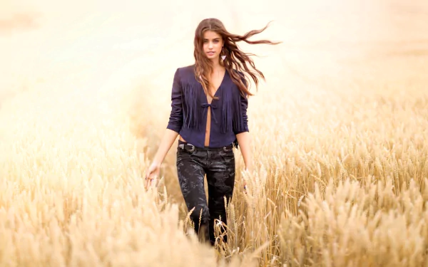 HD desktop wallpaper image featuring a woman, Taylor Marie Hill, walking through a golden wheat field, her hair flowing in the breeze, wearing a dark blue top and jeans.