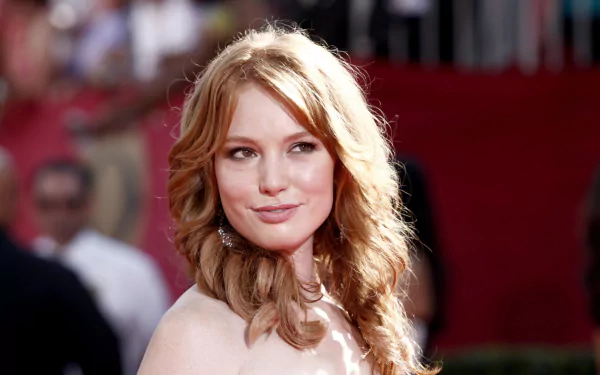 HD desktop wallpaper featuring American actress Alicia Witt with wavy blonde hair, smiling softly against a blurred red carpet background.