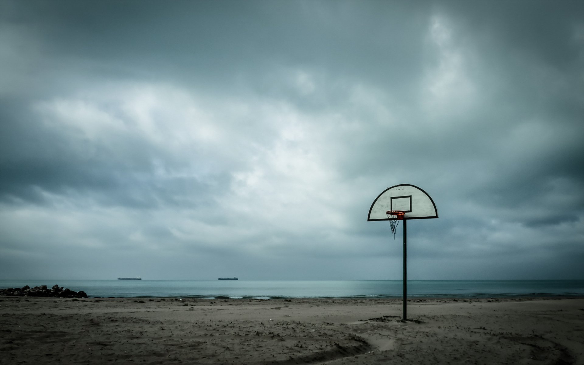 A lonely basketball hoop stands on a sandy beach under a moody, cloudy sky, blending the essence of sports with a tranquil seaside atmosphere. HD desktop wallpaper option.