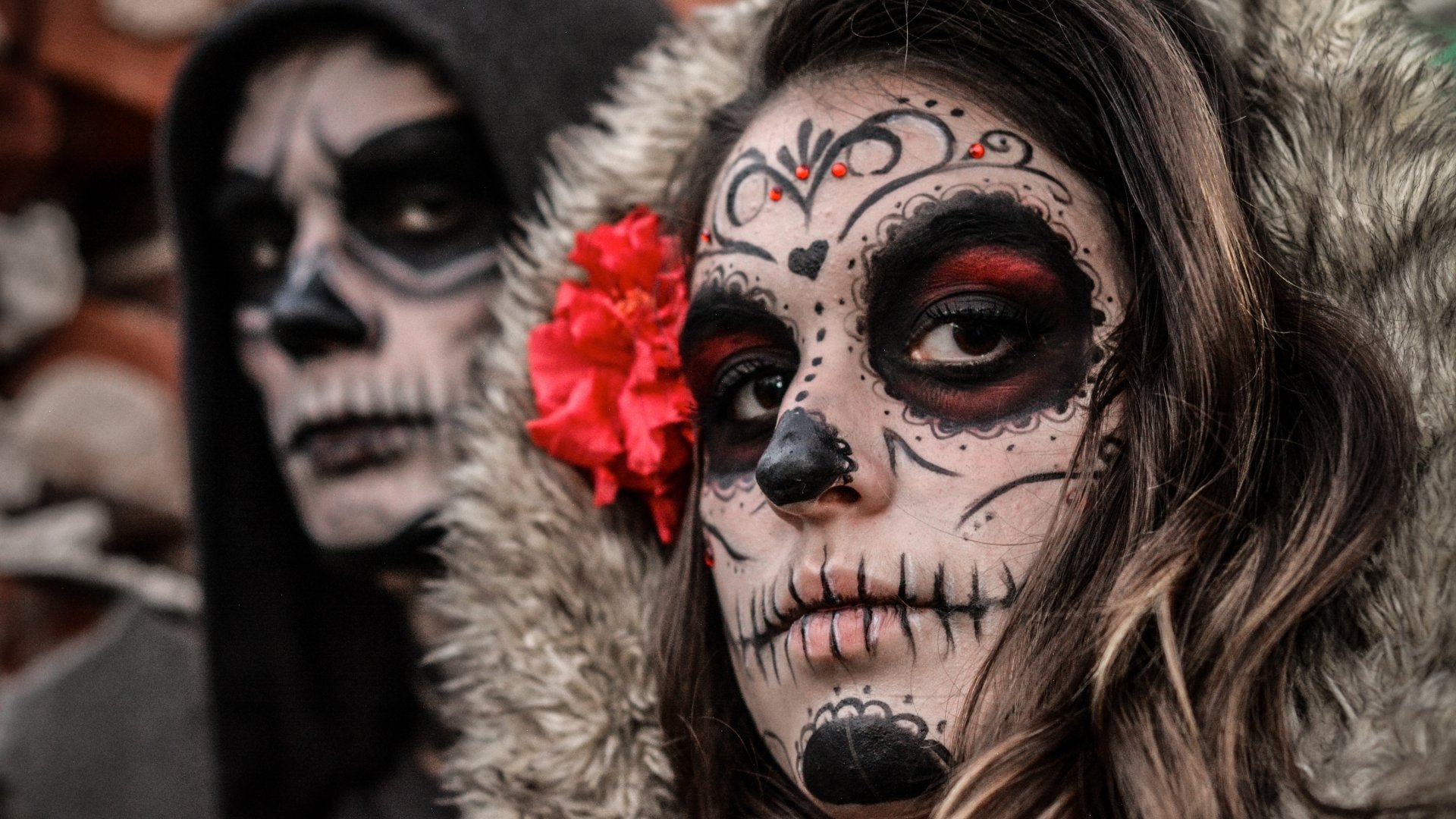 Artistic sugar skull face paint on a woman with a red flower in her hair, captured in stunning 4K Ultra HD for a striking PC desktop wallpaper.