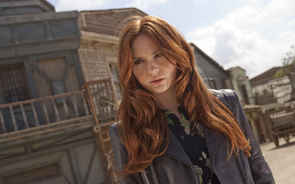 HD desktop wallpaper featuring Scottish actress Karen Gillan with long red hair, standing outdoors against a rustic building backdrop.