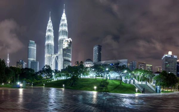HD desktop wallpaper featuring the illuminated Petronas Towers at night in Kuala Lumpur, Malaysia, with surrounding cityscape and lush greenery in the foreground.