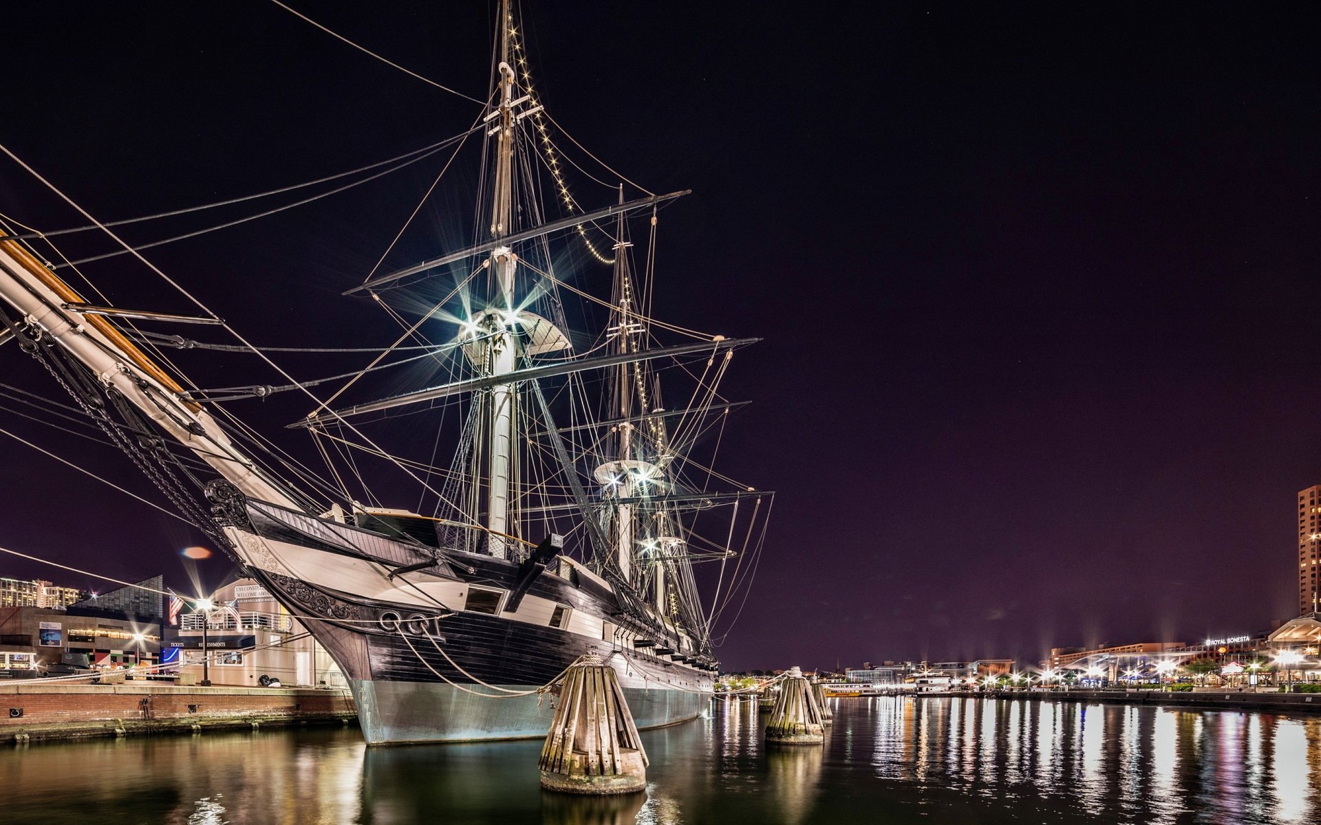 HD desktop wallpaper featuring a historic sailboat docked at night with illuminated rigging and city lights reflecting on calm water.