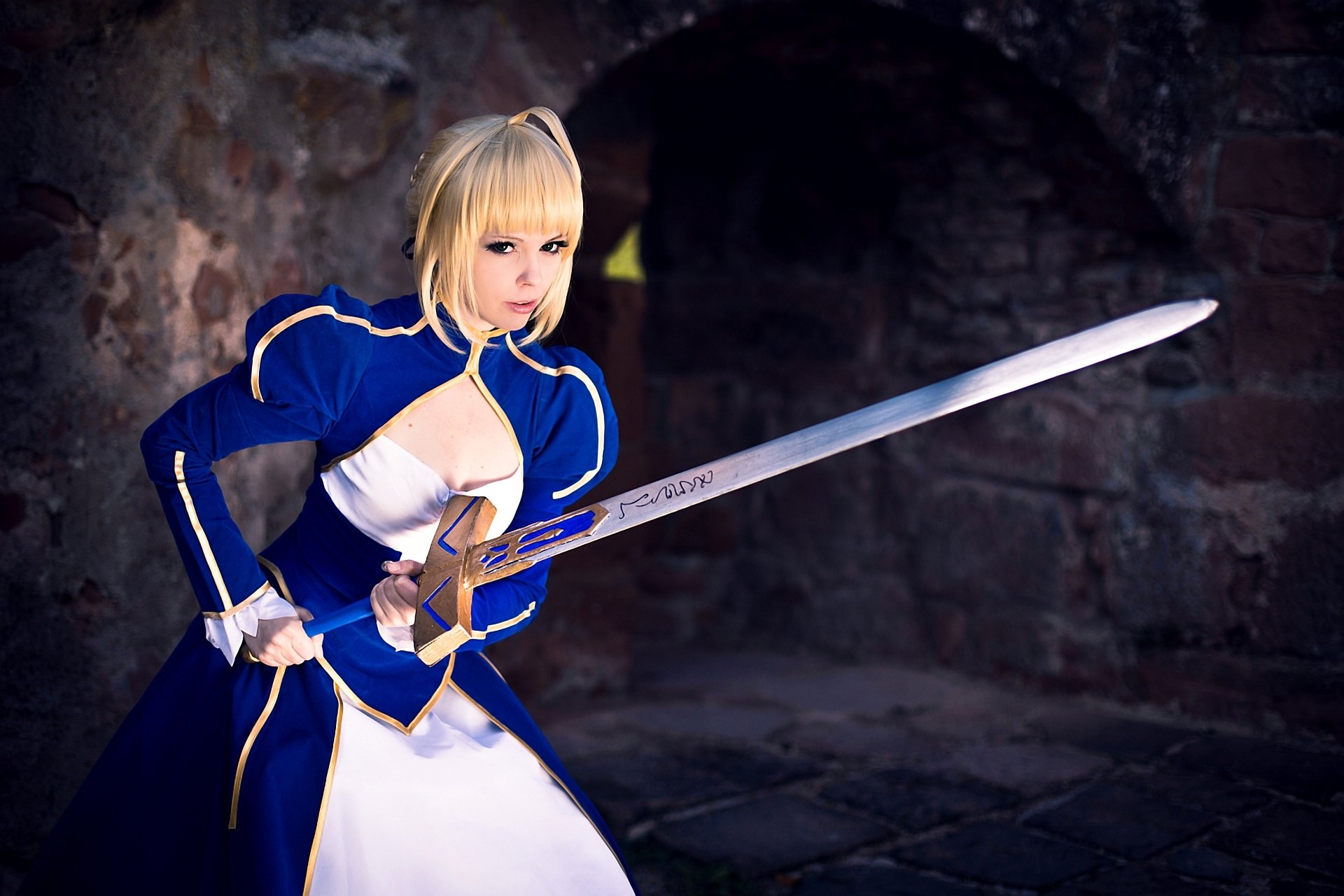 A high-definition wallpaper featuring a woman cosplaying as Saber from the Fate/Stay Night series, poised with a sword in a dramatic setting.
