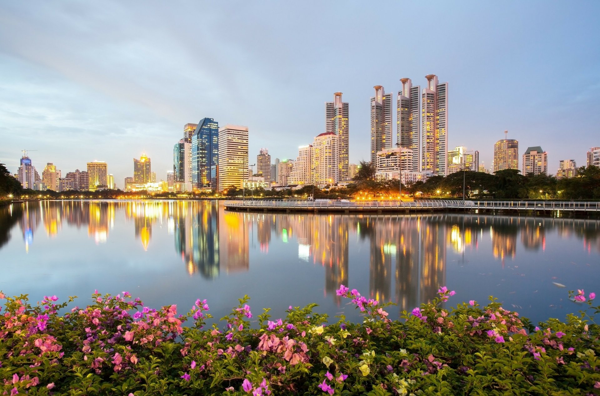 HD desktop wallpaper showing the man-made skyline of Bangkok, Thailand along the tranquil waters of the Chao Phraya River at dusk, with vibrant flowers in the foreground.
