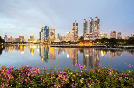 HD desktop wallpaper showing the man-made skyline of Bangkok, Thailand along the tranquil waters of the Chao Phraya River at dusk, with vibrant flowers in the foreground.