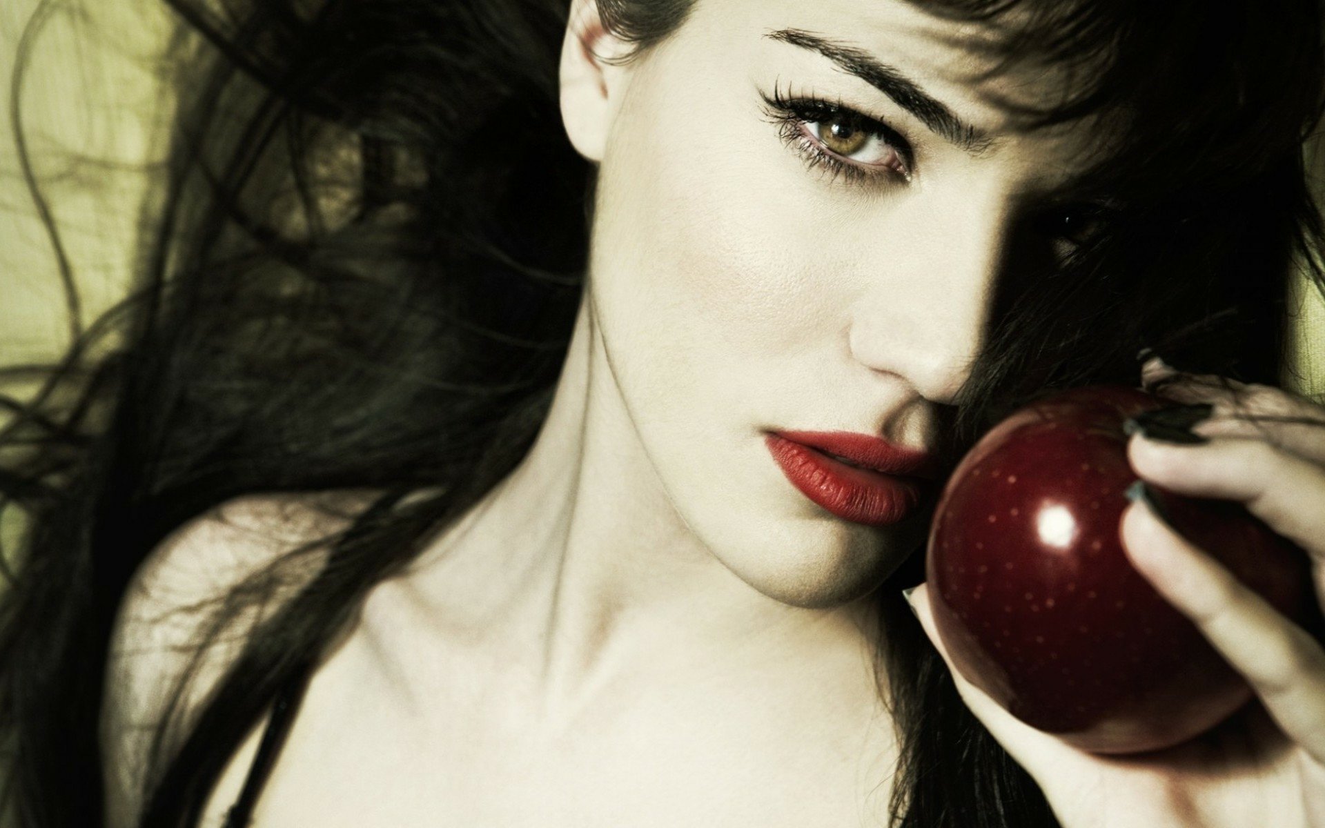 Monica Bellucci gazes captivatingly at the viewer while holding a red apple, framed by flowing dark hair. A striking HD desktop wallpaper that highlights her allure.