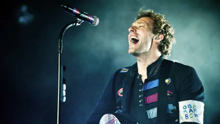A dynamic HD wallpaper featuring a passionate Coldplay performance, showcasing the lead singer singing joyfully into a microphone under dramatic lighting.